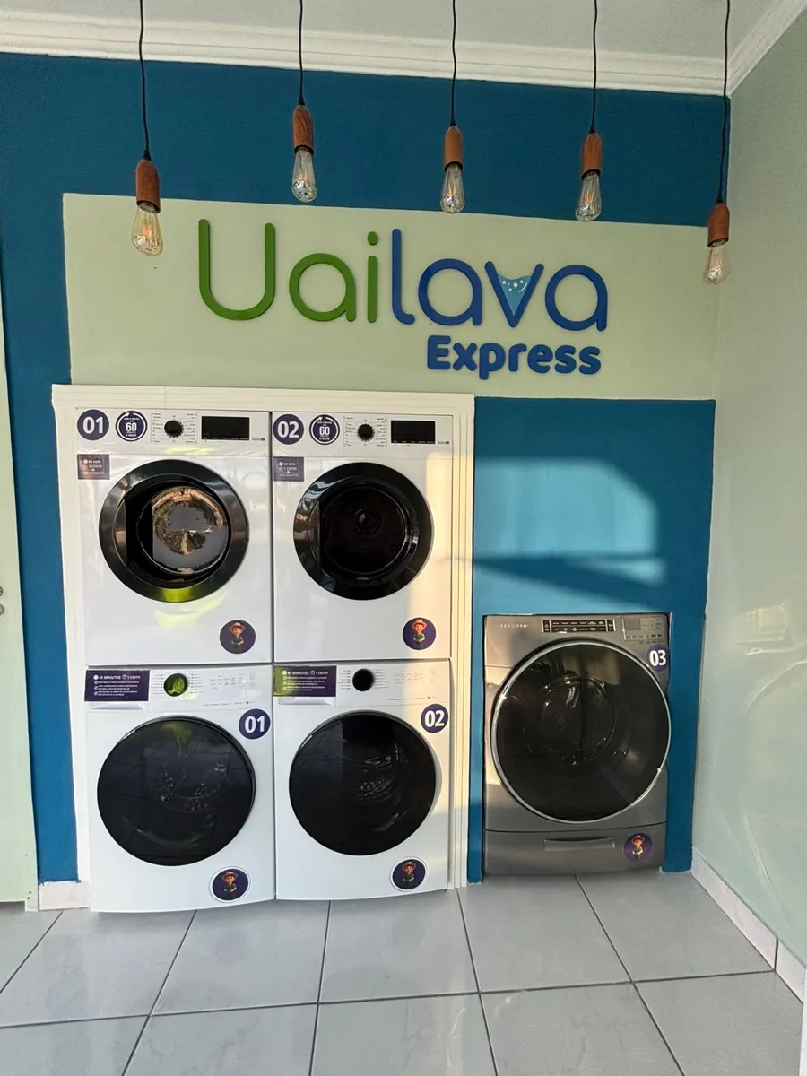 As máquinas da Uailava Express Bragança: duas lavadoras e duas secadoras empilhadas sob o letreiro da marca, e à direita a máquina de 18 kg, com pendentes de filamento acesos.